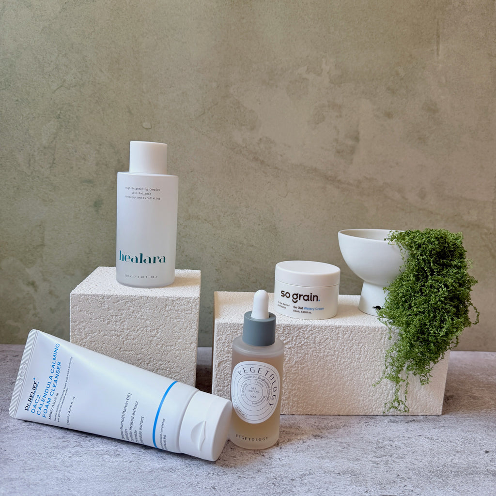 A curated collection of skincare products from the brands Vegetology, Healara, Sograin, and Dr Relief.  styled on textured beige blocks with a small white bowl and trailing green moss against a neutral stone background.