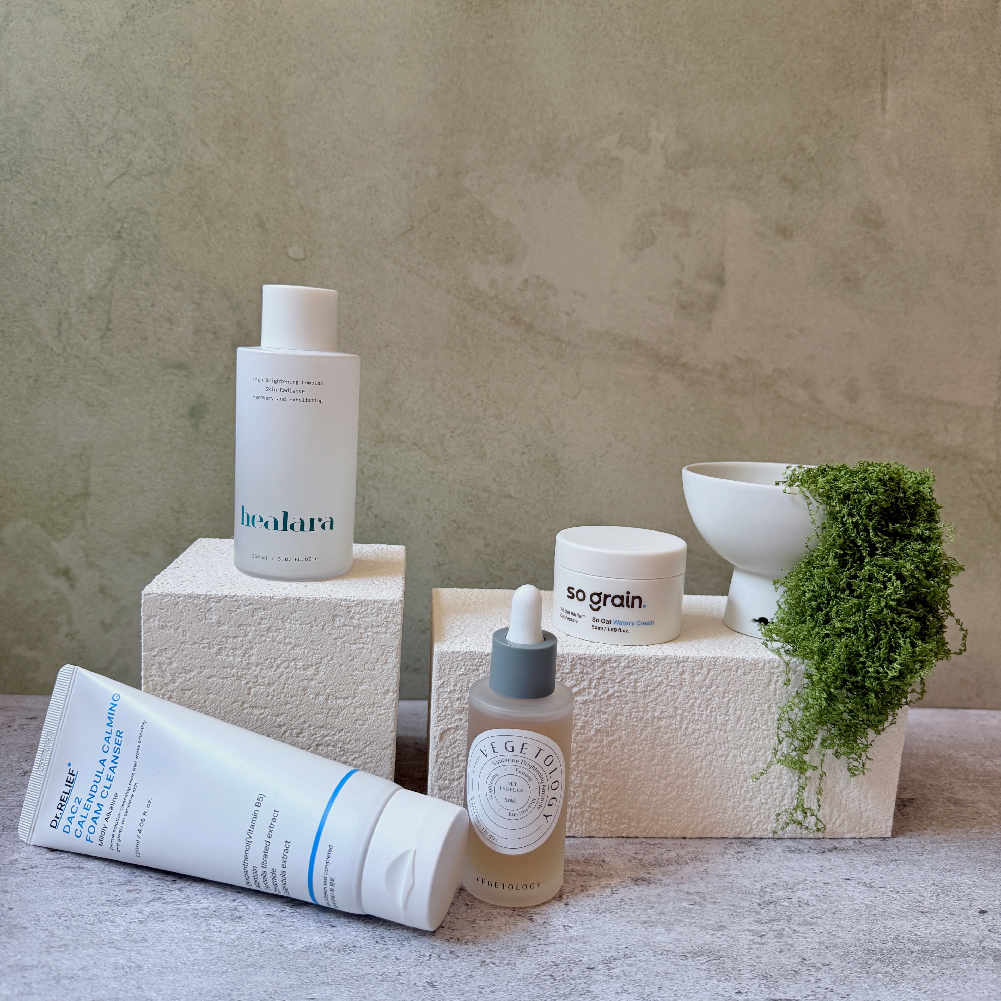 A curated collection of skincare products from the brands Vegetology, Healara, Sograin, and Dr Relief.  styled on textured beige blocks with a small white bowl and trailing green moss against a neutral stone background.